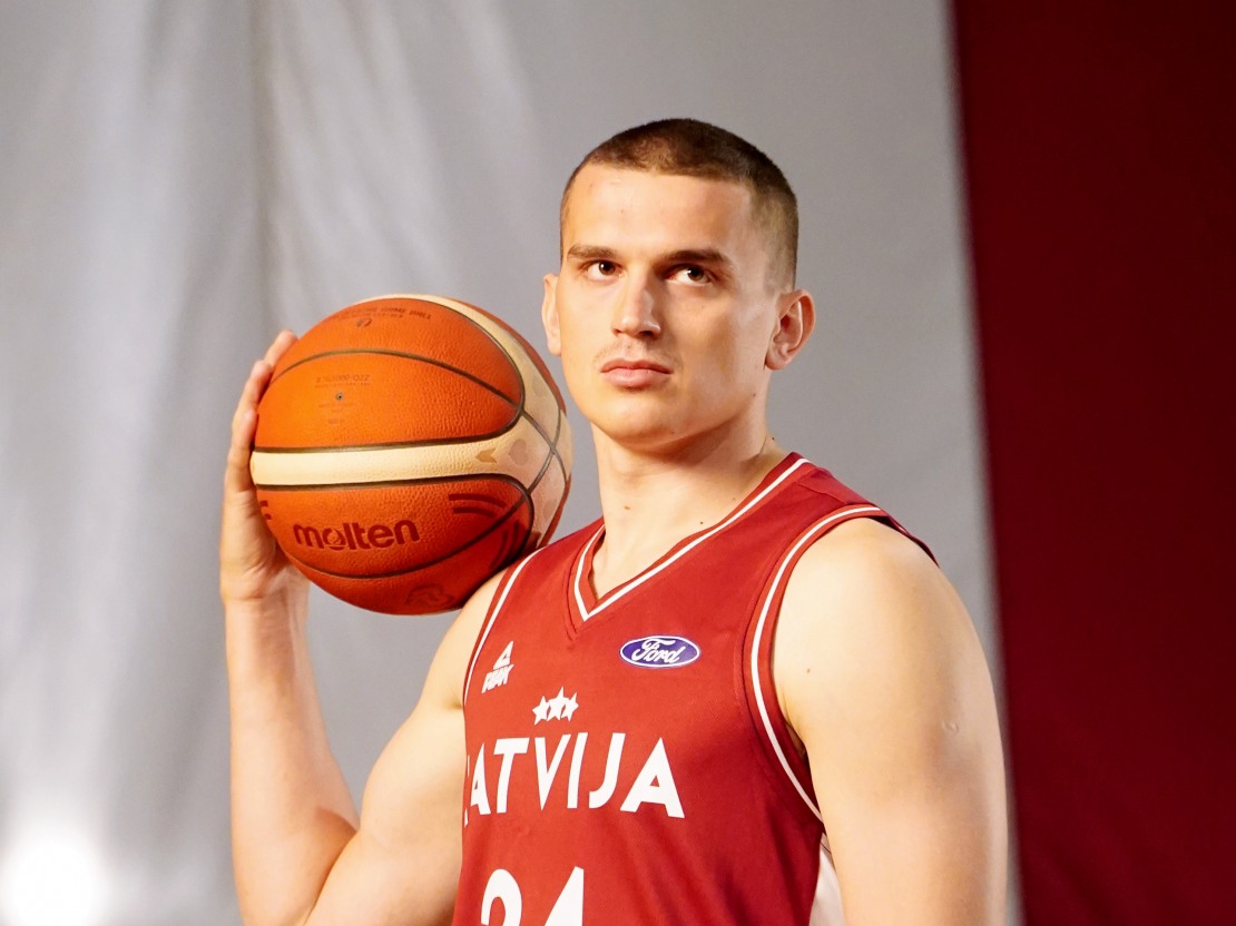 Jauna vīrirša krāsainaios foto. Viņam ļoti īsi mati, sporta sarkanais krekls bez piedurknēm. Uz pleca tur basketbola bumbu.