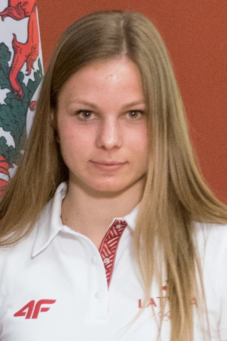 Krāsains jaunas sievietes foto. Viņai gari gaiši mati, baltā blūze ar Latvijas simboliku