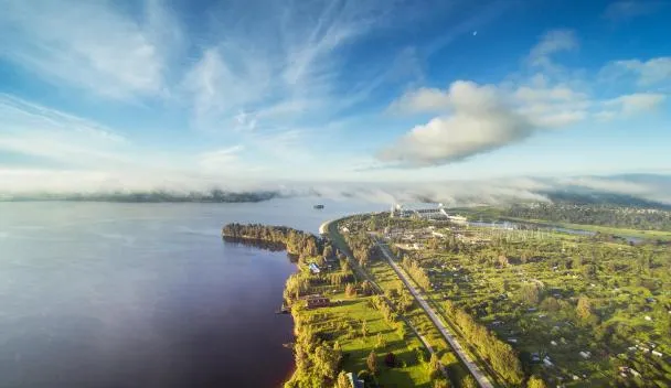 Ainavas skats no putna lidojuma: plaša upe kreisajā pusē, gar tās krastu stiepjas zaļas teritorijas ar ceļu un apdzīvotām vietām. Fonā redzami rūpniecības objekti un tālumā miglas josla, debesīs gaišas mākoņu strēles un zilas debesis.