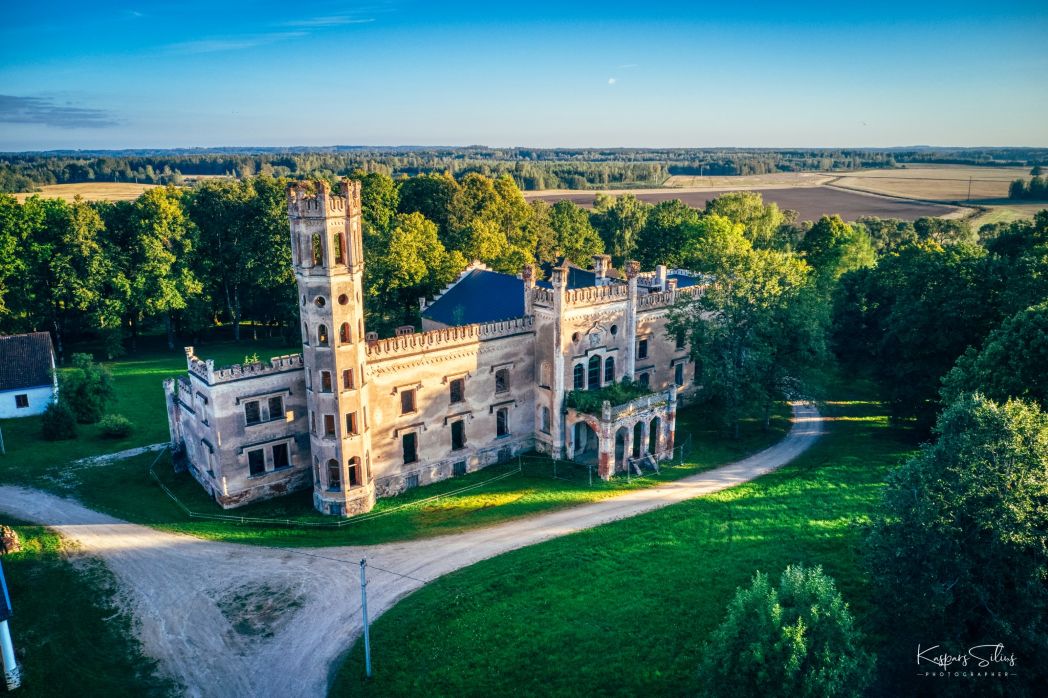 Odzienas muiža – vēsturiska, neogotikas stilā celta ēka ar torni un dekoratīvām arkām, ieskauta koku alejā un lauku ainavā. Skats no augšas.