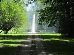 Parka skats ar bruģētu celiņu, kas ved uz strūklaku centrā. Apkārt ir zaļi koki un zālājs, radot mierīgu, dabisku vidi.