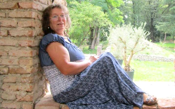 Sieviete, kas sēž pie ķieģeļu sienas ārā, dabas vidē, un skatās kamerā. Sievietei mugurā gara gaiši zila kleita ar īsiem piedurknēm.