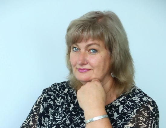 Sievietes portreta foto – viņa skatās kamerā, atbalstot zodu ar roku. Viņai mugurā melni balts puķains tērps.